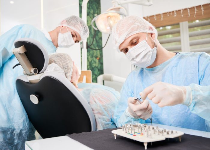 Dentist on foreground, wearing disposable sterile clothes, putting surgical nozzle on drill before implant installation. Patient and assistant on blurred background. Concept of implant placement.